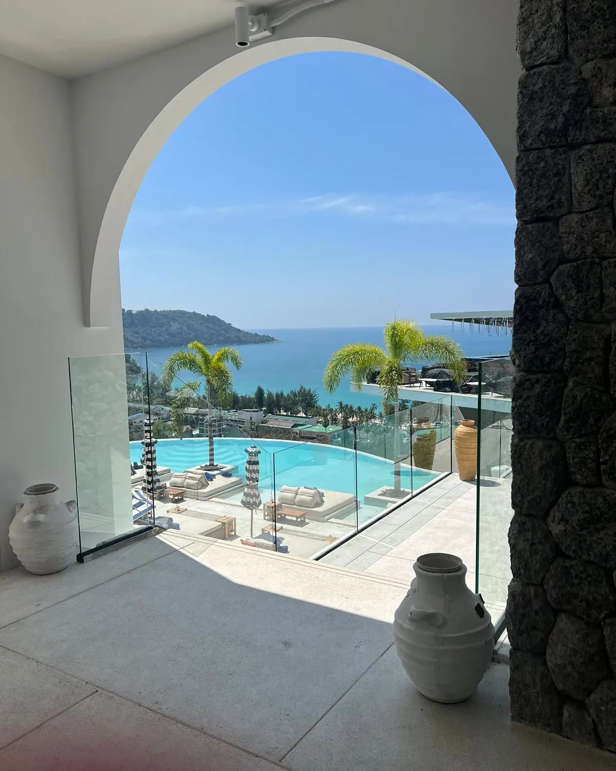 Piscine à débordement et mer d'Andaman vues depuis l'arche blanche d'une villa de Phuket, palmiers et transats au bord du bassin
